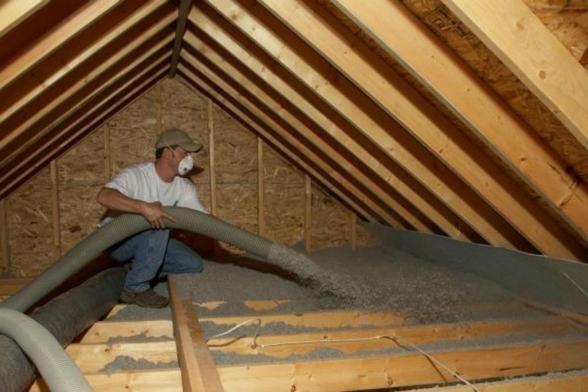 A worker in a protective mask uses a large hose to install insulation in an attic with exposed wooden beams, focusing on the task with care.