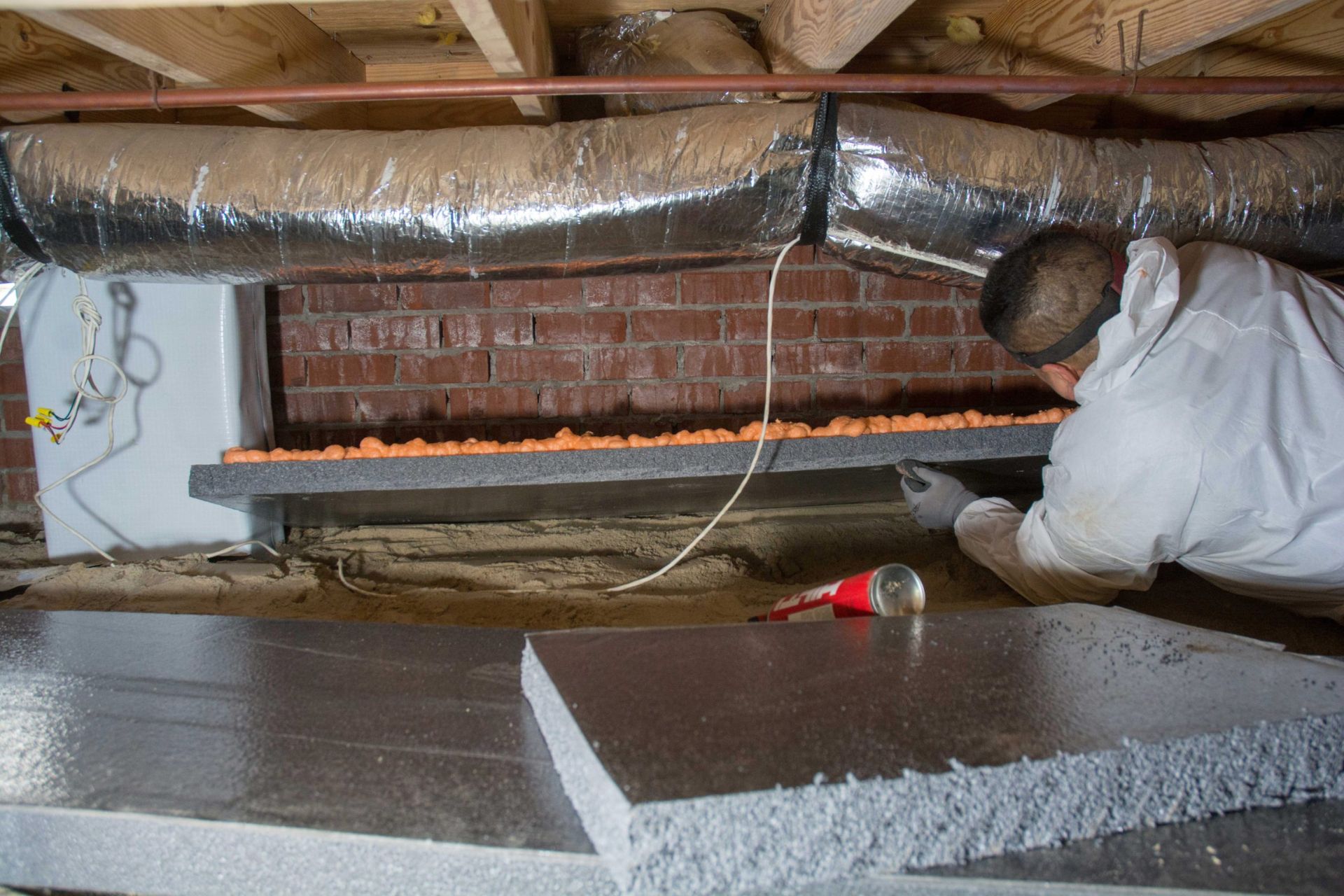 A man in a white coat installs insulation in a crawl space, handling foam panels near HVAC ducts against a brick wall.