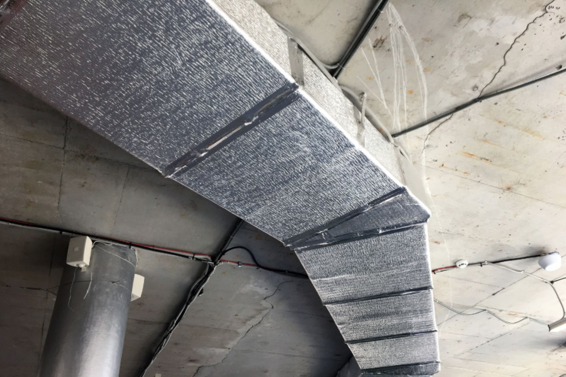 Silver ductwork running along a concrete ceiling, with exposed wiring and pipes visible in a commercial or industrial setting.