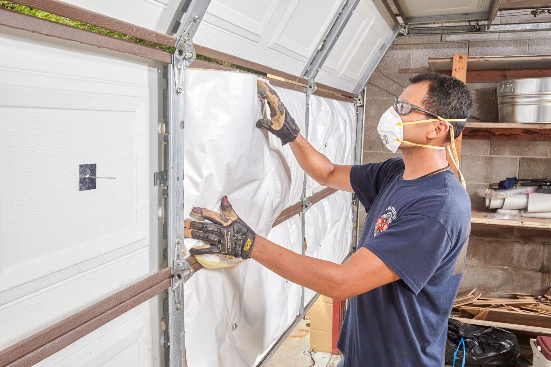 A man in a blue shirt and gloves installs insulation on a garage door in a workshop, with tools scattered in the background.