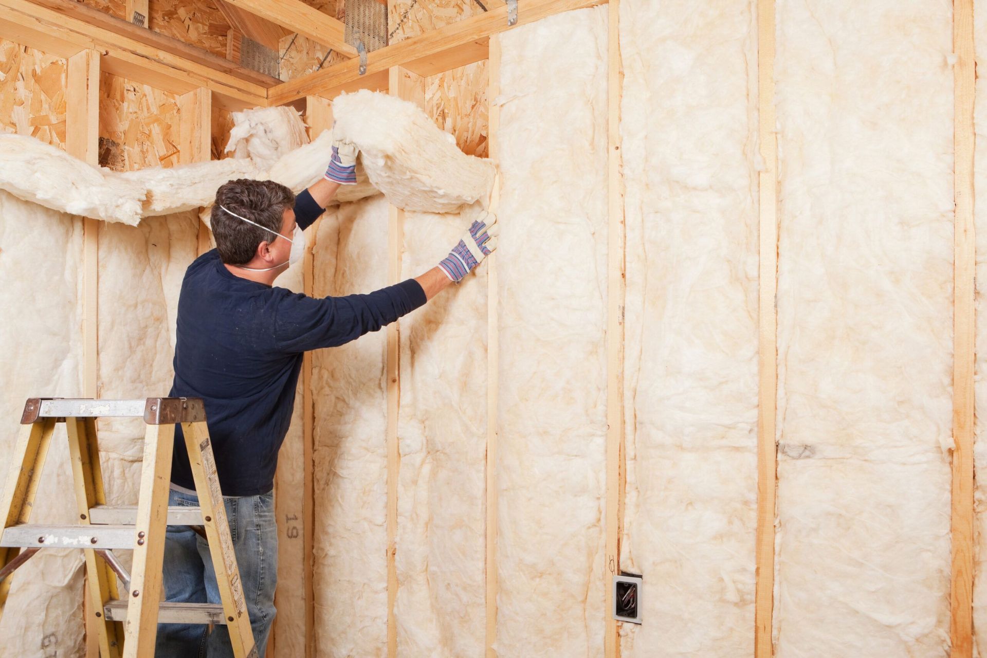 A man in gloves installs insulation between wooden studs in a house under construction, standing on a ladder.