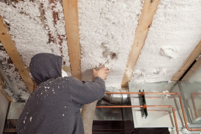 A man in a hoodie installs insulation on a ceiling, focused on his work in a well-lit room.