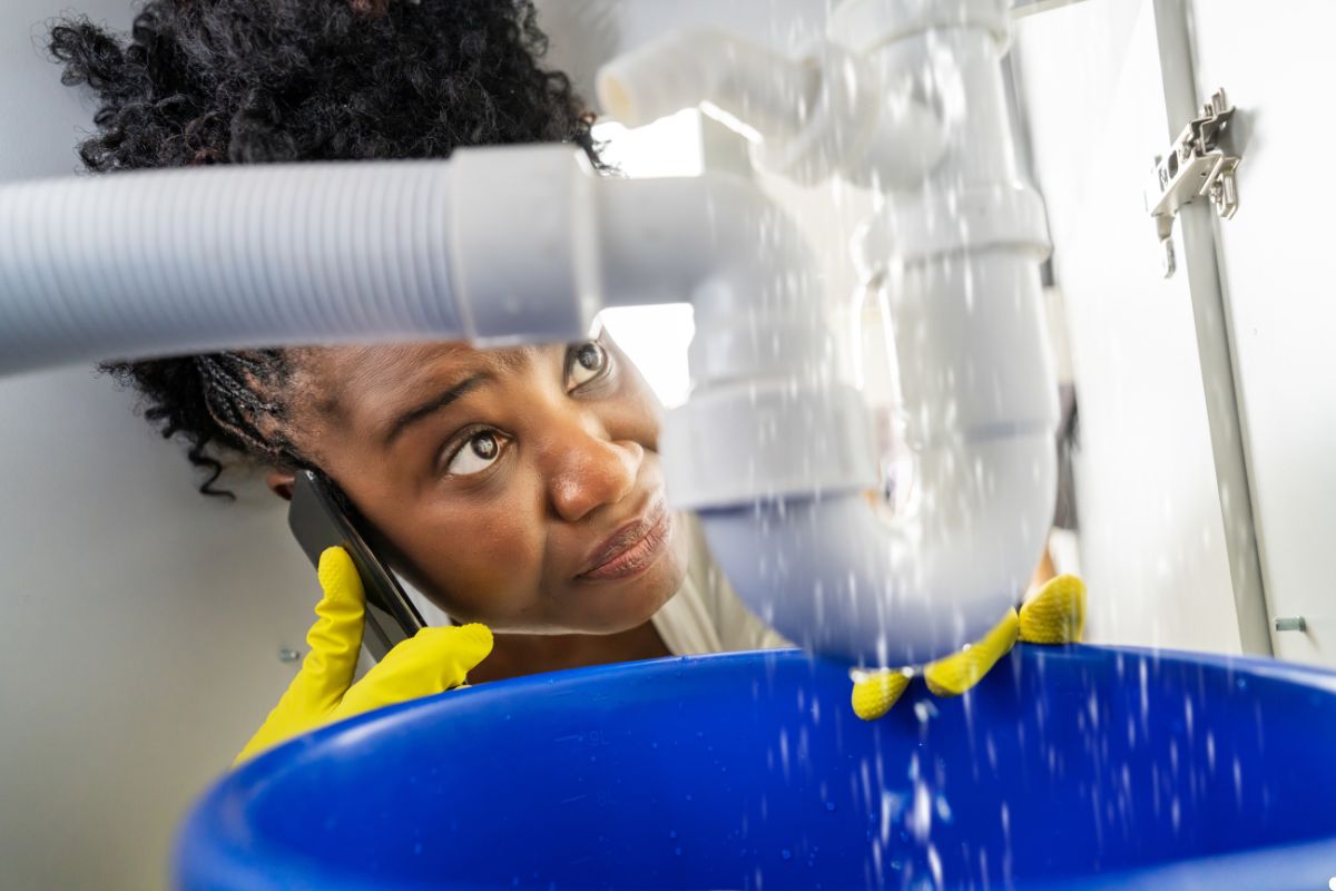 A woman holds a phone beneath a sink, appearing to check for plumbing issues or communicate with someone.